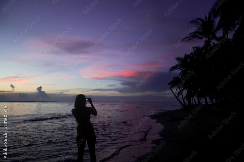 Silhouette of solo female backpacker takes a photo of a sunset using her phone on a paradise beach in Central America