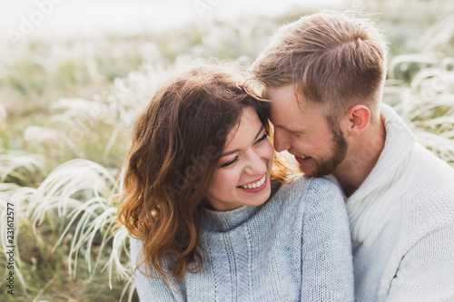 Happy young loving couple sitting in feather grass meadow, laughing and hugging, casual style sweater and jeans