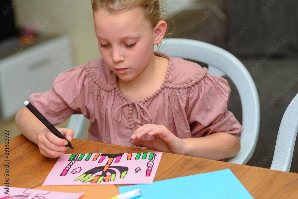 Needlework, crafts children.Little girl create a greeting card image of the Jewish holiday of Hanukkah. Kid pastes stickers - candles and drawing menorah, dreidel, oil jar on paper on wooden table. Needlework, crafts children.Little girl create a greeting card image of the Jewish holiday of Hanukkah. Kid pastes stickers - candles and drawing menorah, dreidel, oil jar on paper on wooden table.