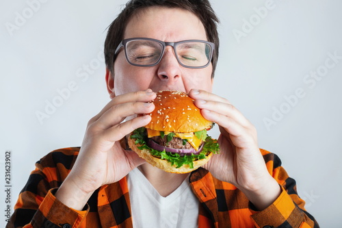 A young guy with glasses holding a fresh Burger. A very hungry student eats fast food. Hot helpful food. The concept of gluttony and unhealthy diet. With copy space for text