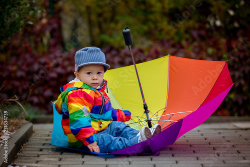 Sweet funny child with rainboy coat and multicolored umbrella jumping on puddles iand playing outdoors photo