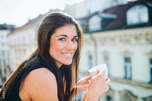 Young businesswoman with cup of coffee standing on a terrace in an office in city.