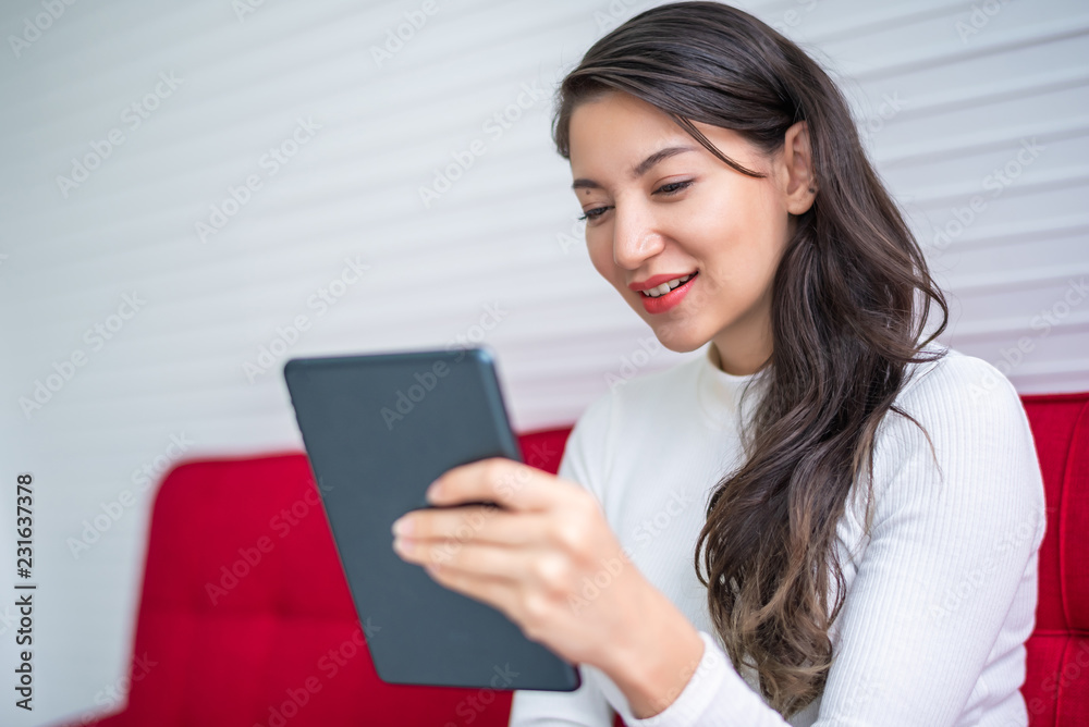 Beautiful attractive young Asian woman using smart phone or cellphone or tablet while sitting on red sofa at living room in the morning Beautiful attractive young Asian woman using smart phone or cellphone or tablet while sitting on red sofa at living room in the morning