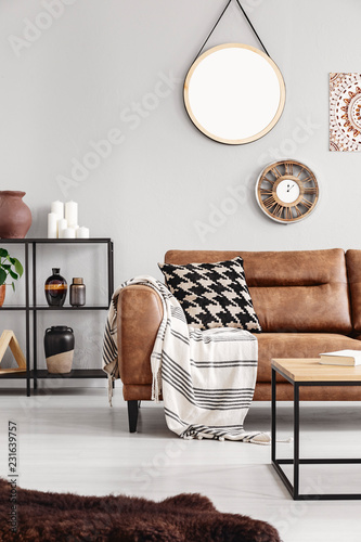 Vertical view of warm ethno living room with leather couch with patterned pillow and mirror and clock on beige wall, real photo