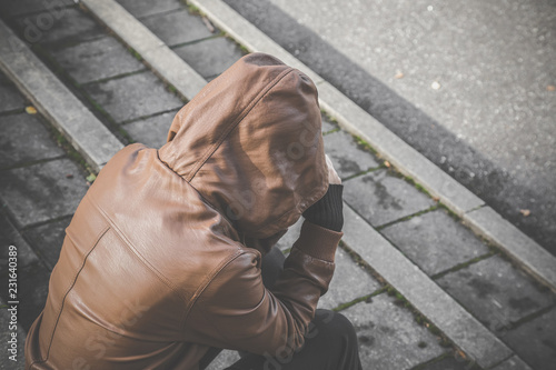 Young person in brown leather jacket with hood sitting on stairs and suffering from life difficulties, broken heart or psychological problems. Dark, gloomy thoughts. Social isolation. photo