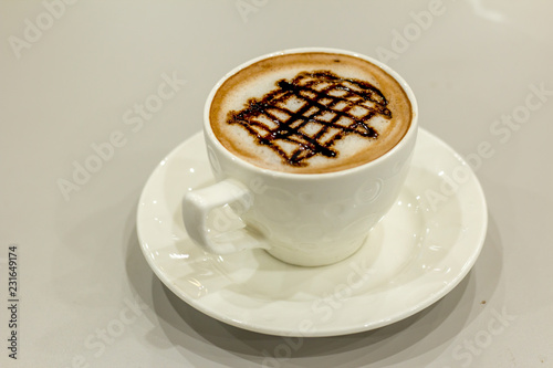 The white cup of hot capuchino and chocolate syrup on top on the wooden table background.
