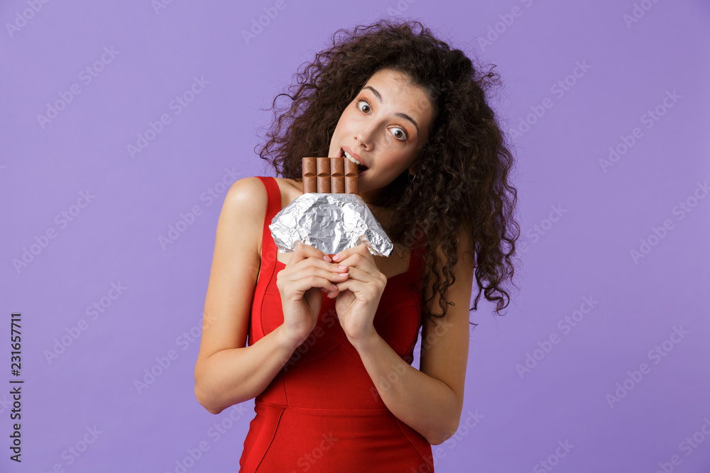 Image of charming woman 20s wearing red dress smiling and eating chocolate, standing isolated over violet background Image of charming woman 20s wearing red dress smiling and eating chocolate, standing isolated over violet background