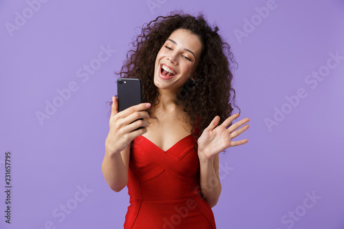 Image of joyous woman 20s wearing red dress using black smartphone, standing isolated over violet background