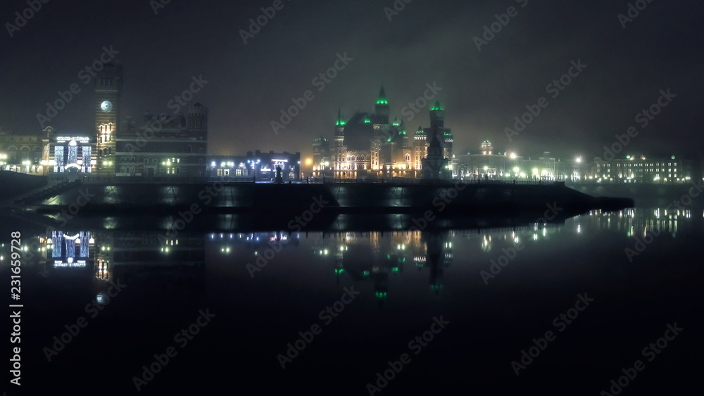 mystical towers and castles of the night city illuminated by lights in ominous fog are reflected symmetrically in the cold river
