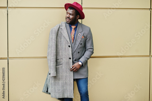 Stylish African American man model in gray coat, jacket tie and red hat posed against golden background. photo