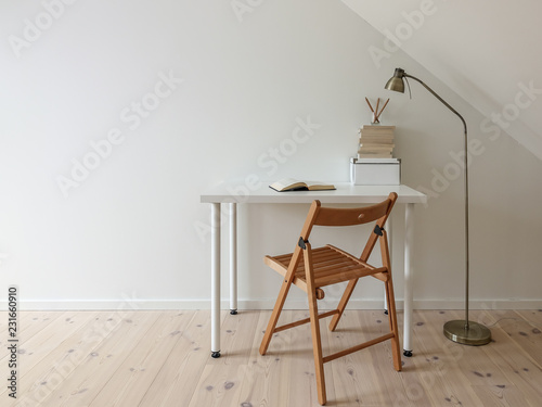 Simple workspace interior. Open book in minimalist home office room. Lamp, books, wooden floor and folding chair. Empty white wall and desk for copy space.