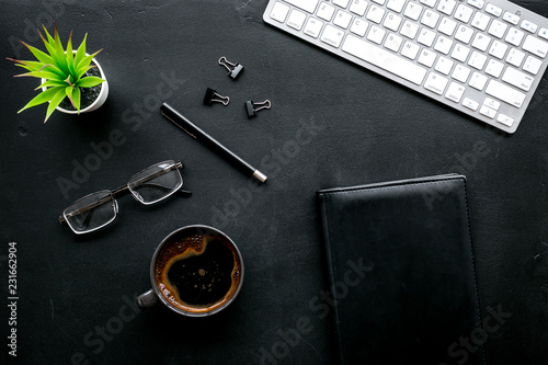 Black strict office desk, monochrome. Computer keyboard, expensive black notebook, glasses, coffee. Top view