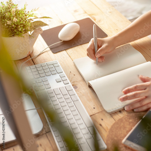 Hands of a Business woman is writing book as notepad or Take notes on Her wooden table desk  . Business Success Working at home office with notebook and computer for marketing and part time Concept.