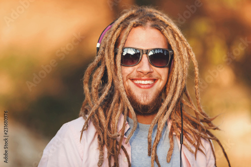 Portrait of hipster blonde bearded boy with dreads wearing headphones and glasses walking and smiling outdoors an the sunny autumn day
