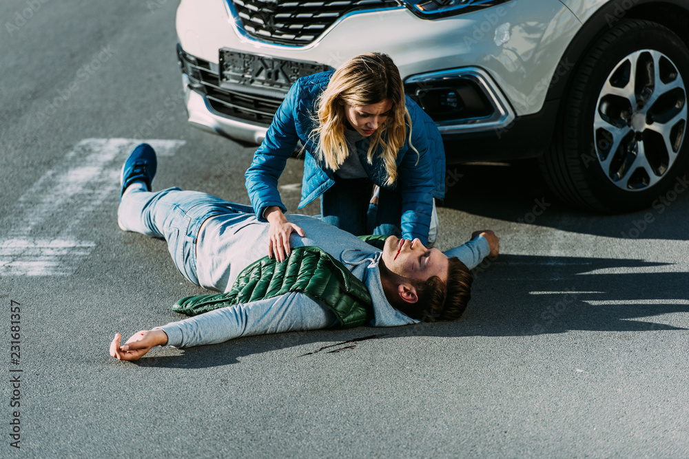 high angle view of scared young woman touching injured man lying on road after car accident - obrazy, fototapety, plakaty 