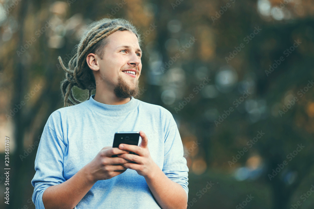 Hipster blonde bearded man with dreads wearing casual clothes typing messages on his smartphone and looking on the nature views, outdoor view Hipster blonde bearded man with dreads wearing casual clothes typing messages on his smartphone and looking on the nature views, outdoor view