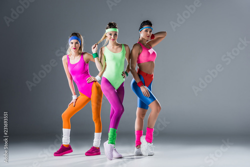 Fototapeta Naklejka Na Ścianę i Meble -  attractive sporty girls in 80s style sportswear posing together and looking at camera on grey