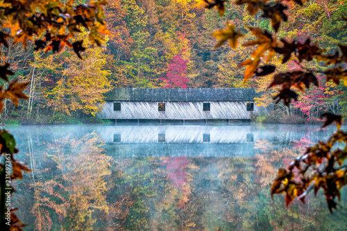 Covered bridge in the fall, As mist rises from the calm waters, fall colors are reflected around the covered bridge.