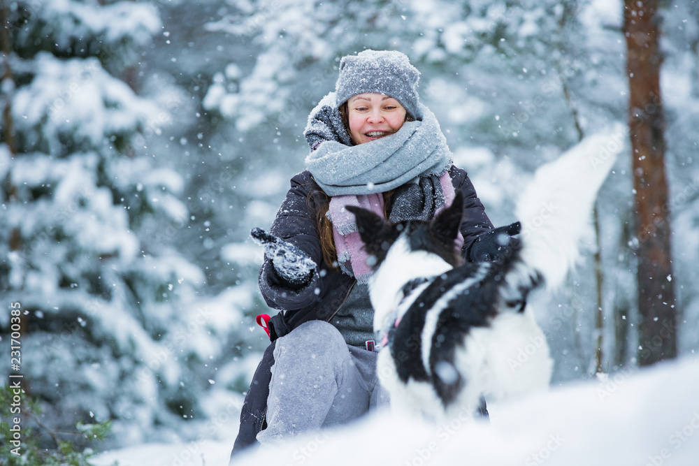 Woman playing with dog in snowy forest. Running and jumping happy pet, girl laughing, having fun. Beautiful winter landscape with trees in snow. 