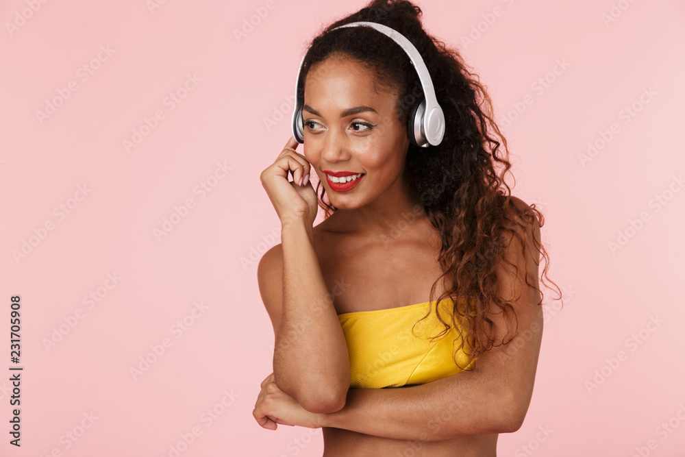 Fototapeta premium Woman in swimwear posing isolated over pink background listening music with headphones.