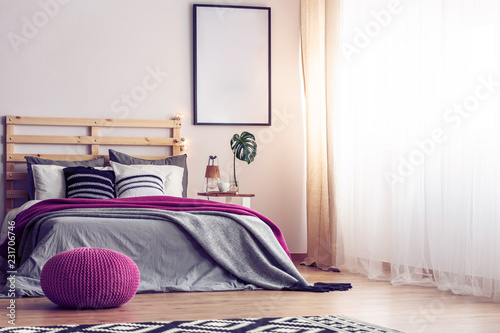 Mockup in black frame on empty wall of bright scandinavian bedroom with comfortable bed with grey bedding, pink blanket and pouf, real photo with copy space lam