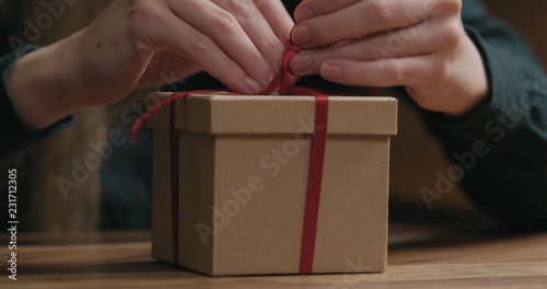 Closeup shot of female hands tying red ribbon bow on craft paper gift box