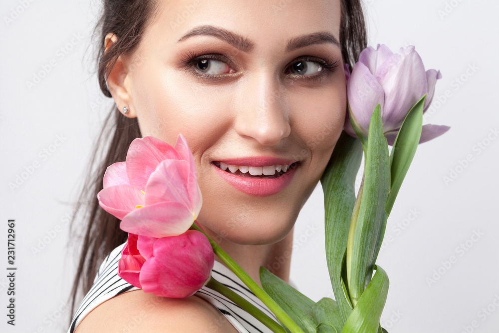 closeup ortrait of young brunette beautiful woman with makeup in striped dress, standing and holding tulips and looking away with toothy smile. indoor studio shot, isolated on light grey background. closeup ortrait of young brunette beautiful woman with makeup in striped dress, standing and holding tulips and looking away with toothy smile. indoor studio shot, isolated on light grey background.