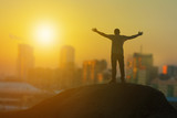Man businessman on top of a mountain on the background of the city in the rays of sunset. Raising his hands up. Business concept idea, success and achievement, happy, career.