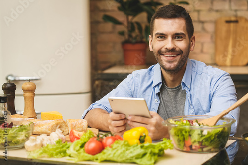Man preparing delicious and healthy food in the home kitchen on a sunny day. Using tablet computer for searching recipes.