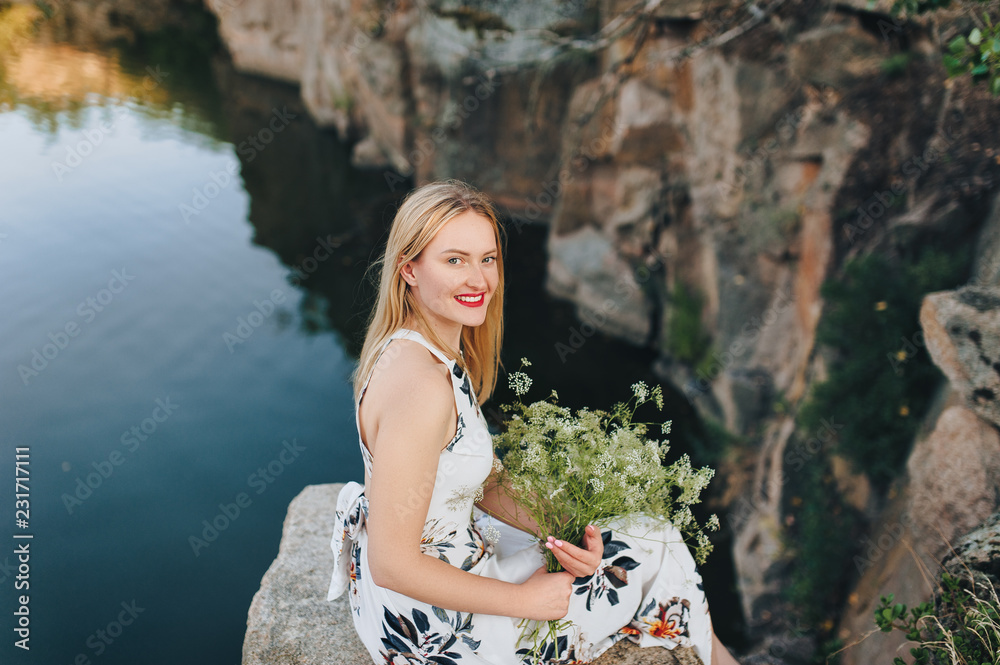 Beautiful blonde girl sitting on a cliff with a bouquet on the background of the lake and stones. Waiting for a sweetheart. Portrait of the bride. Evening relaxation in nature. Beautiful blonde girl sitting on a cliff with a bouquet on the background of the lake and stones. Waiting for a sweetheart. Portrait of the bride. Evening relaxation in nature.