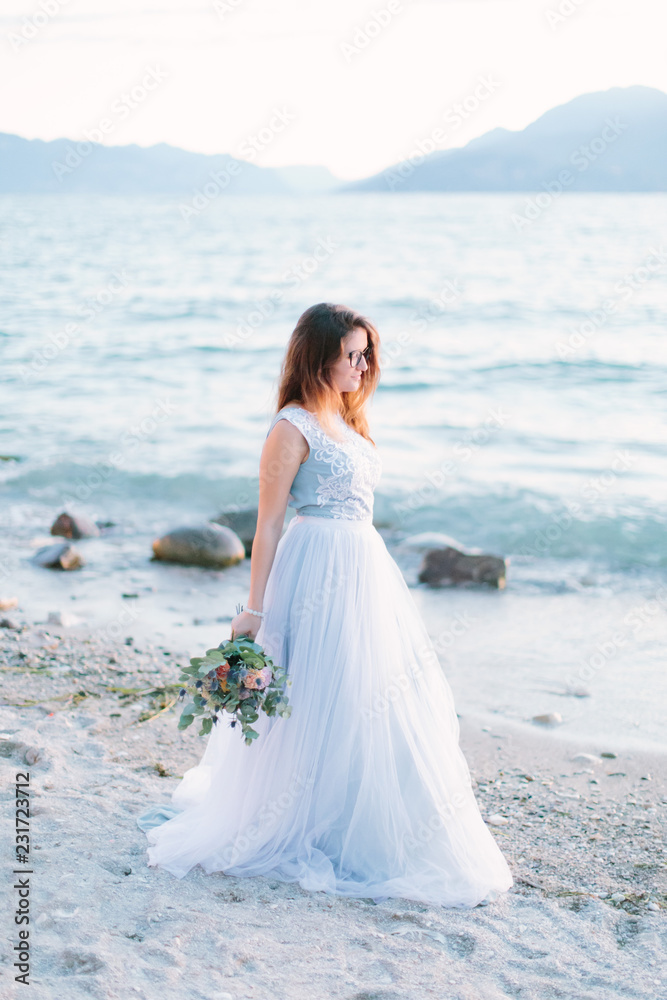 Beautiful bride in blue elegant dress holds a bouquet of flowers walking on the shores of Lake Garda. Sirmione, Italy