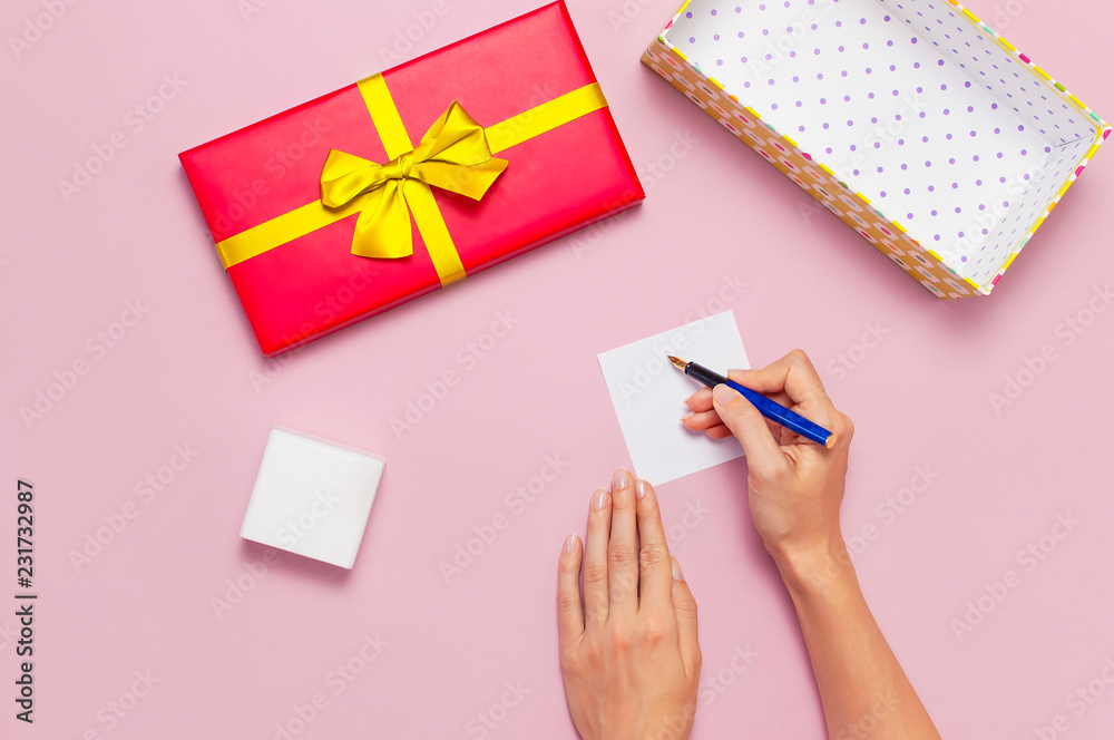Flat lay top view womens hands write congratulations on empty sheet, red present box with gold ribbon on light pink background. Holiday concept birthday gift Congratulations background copy space Flat lay top view womens hands write congratulations on empty sheet, red present box with gold ribbon on light pink background. Holiday concept birthday gift Congratulations background copy space