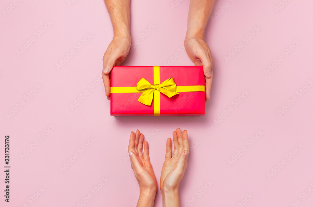 Φωτογραφία Top view of male and female hands holding red gift box with golden ribbon on pink background Flat lay
