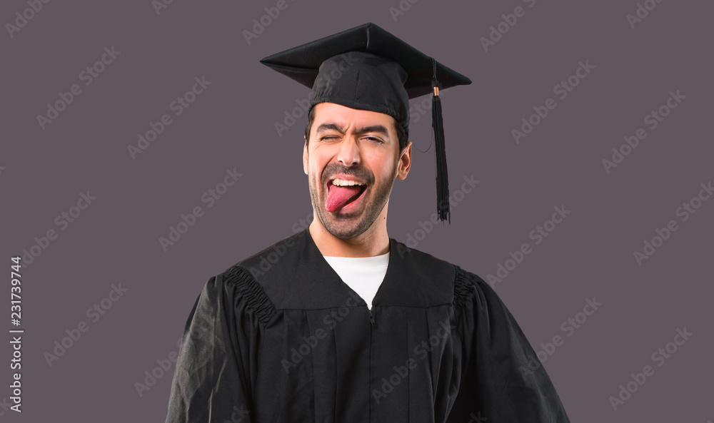 Man on his graduation day University showing tongue at the camera having funny look on violet background