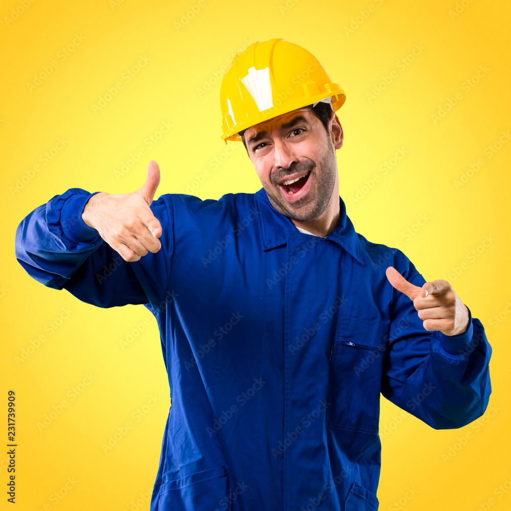 Young workman with helmet pointing with finger at someone and laughing a lot on yellow background Young workman with helmet pointing with finger at someone and laughing a lot on yellow background