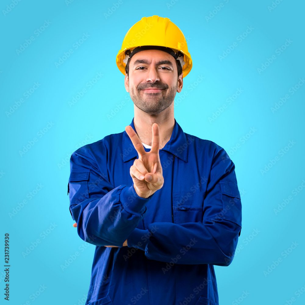 Young workman with helmet smiling and showing victory sign on blue background Young workman with helmet smiling and showing victory sign on blue background