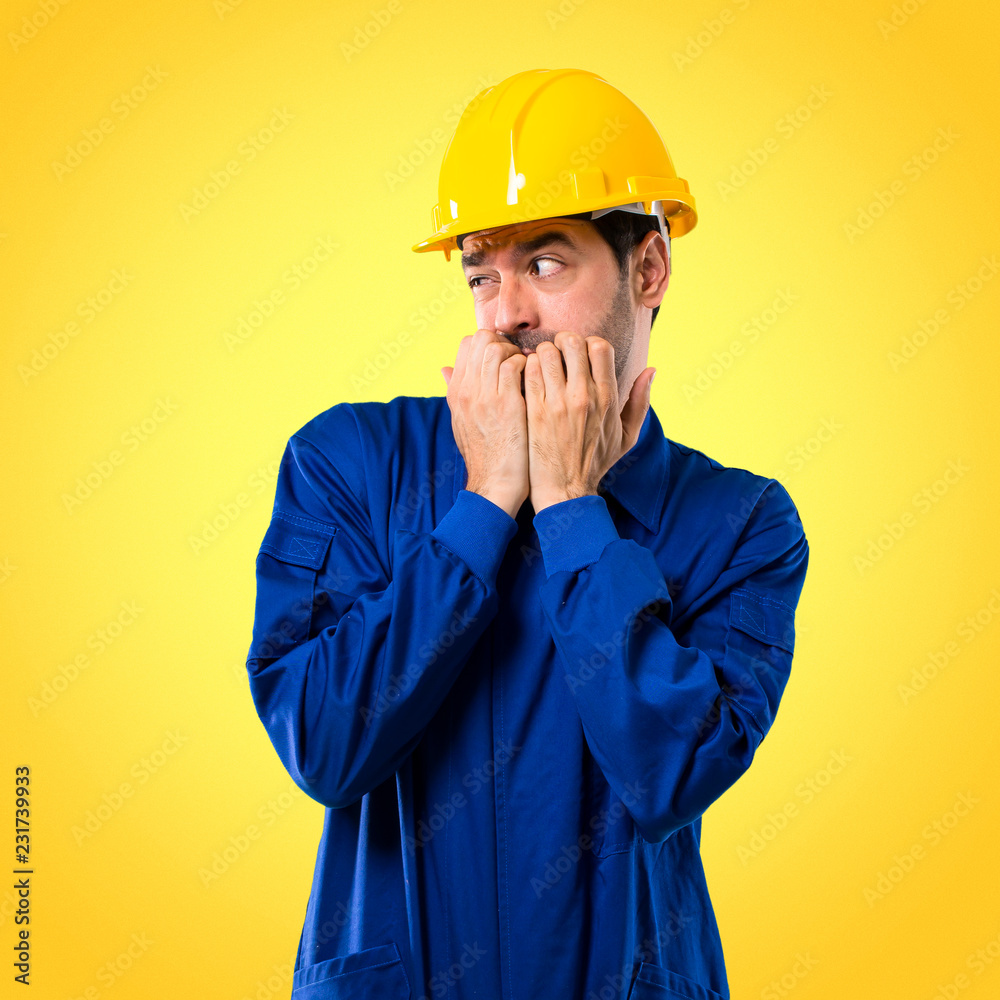 Young workman with helmet is a little bit nervous and scared putting hands to mouth on yellow background
