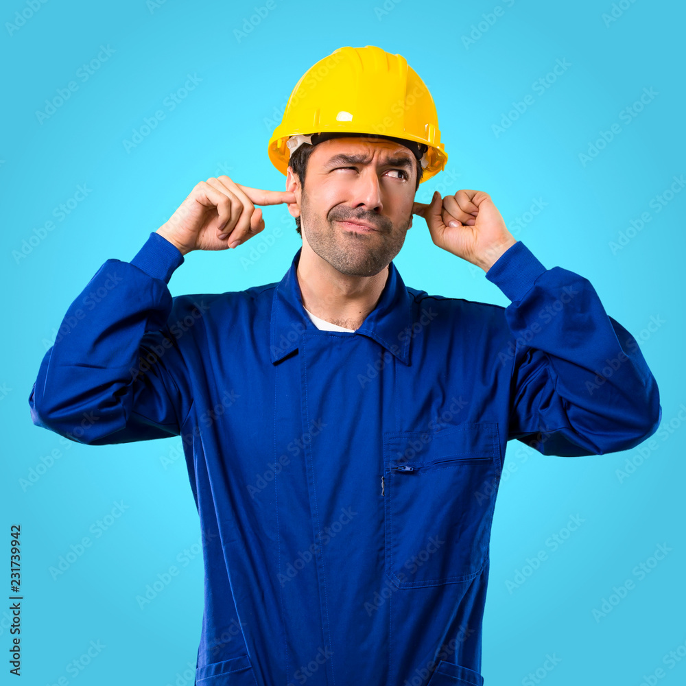Young workman with helmet covering both ears with hands on blue background
