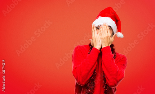 Man with red clothes celebrating the Christmas holidays covering eyes by hands and looking through the fingers on red background