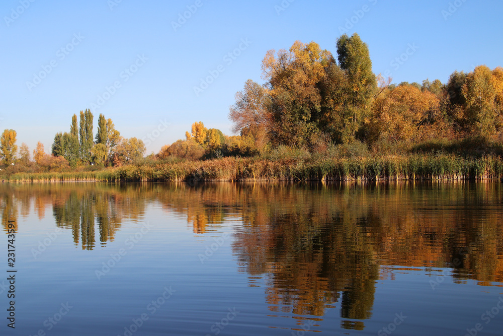 Beautiful pacifying landscape outside the city. The calm water of the lake reflects the colorful autumn trees. Bright colors of autumn. Seasons. November. Natural photo background Beautiful pacifying landscape outside the city. The calm water of the lake reflects the colorful autumn trees. Bright colors of autumn. Seasons. November. Natural photo background