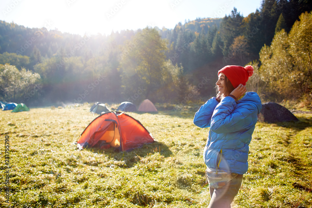 happy smiling woman in a blue down jacket is putting on a red cap. Woman is standing in campsite on a meadow in the autumn forest on a bright sunny foggy morning - obrazy, fototapety, plakaty 