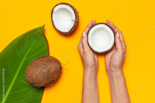 Ripe coconut in female hands and tropical leaves on yellow colored background minimal flat lay top view. Pop art design creative summer, food concept. Tropical fruit whole and half abstract background