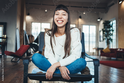 Portrait of nice asian woman sitting in leather chair in big loft apartment and smiling