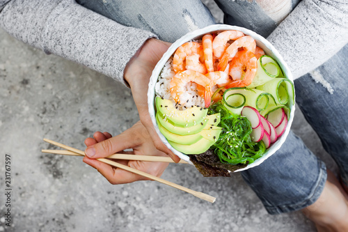 Girl in jeans holding shrimp poke bowl with seaweed, avocado, cucumber, radish, sesame seeds.