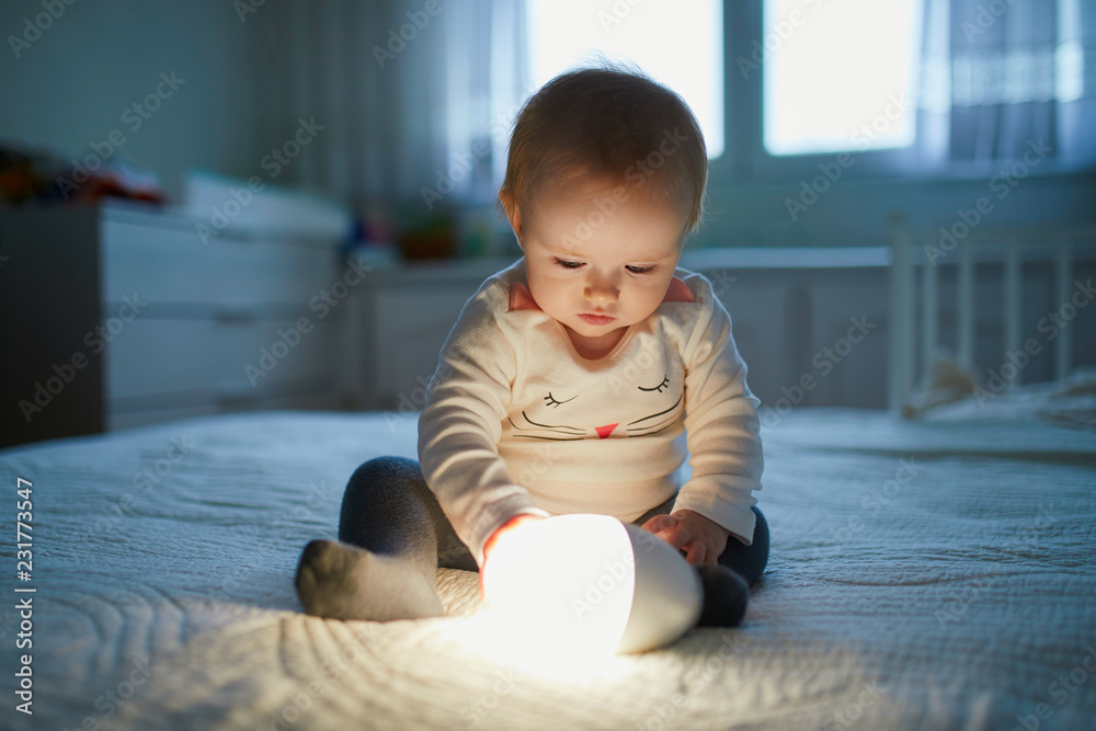 Adorable baby girl playing with bedside lamp in nursery - obrazy, fototapety, plakaty 