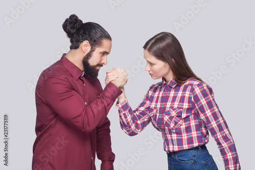 Profile side view portrait of bearded man and woman in casual style standing in armwrestling pose and looking at each other with serious face. indoor studio shot, isolated on gray background.