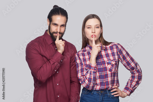 Portrait of serious bearded man and woman in casual style standing and looking at camera with fingers on lips and showing silent sign. indoor studio shot, isolated on gray background.