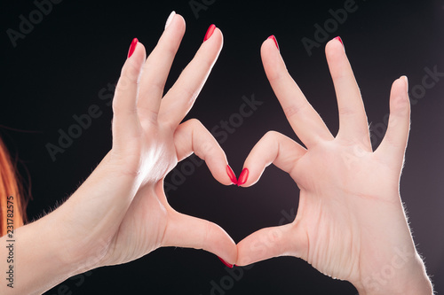 Crop of two hadns together showing heart by fingers and gesturing symbol of amour. Isolated love sign making by hands with red manicured nails on black studio background.  photo