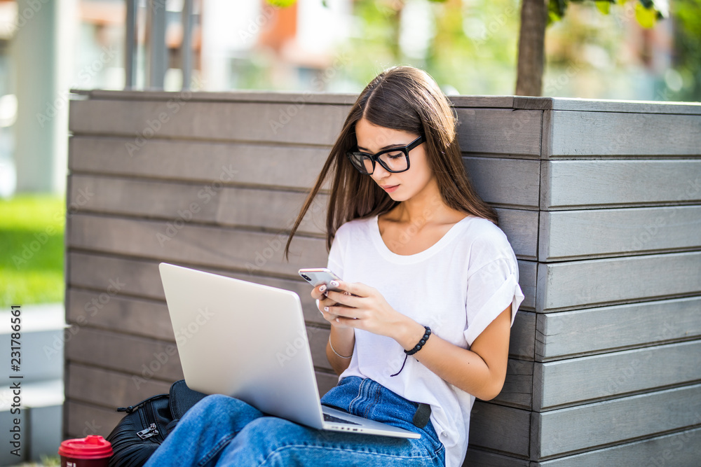 Pretty young woman sitting on bench in city on summer day and reading text message on cell phone while using silver laptop and drinking takeaway coffee Pretty young woman sitting on bench in city on summer day and reading text message on cell phone while using silver laptop and drinking takeaway coffee