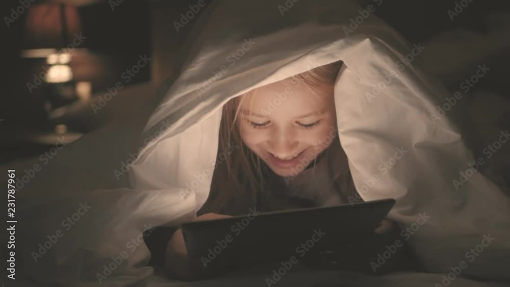 Teenage girl in bed playing a tablet in social internet in the dark light. Close up of little girl watching video on the digital tablet at night. Teenage girl in bed playing a tablet in social internet in the dark light. Close up of little girl watching video on the digital tablet at night.